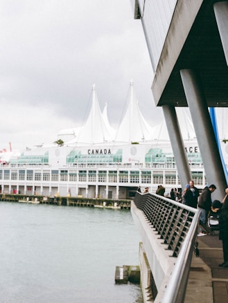 A waterfront scene featuring a modern building with large glass windows and white peaked roofs. People walk along the railing of a promenade that overlooks the water. The word 'CANADA' is marked prominently on the building, and the structure appears to be part of a large complex, likely a convention center.