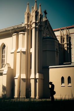 A gothic-style building with tall, ornate spires and arched windows. The structure is lit by warm sunlight, creating contrasting shadows. In the foreground, a silhouette of a person with a hat and a bag is visible, adding a narrative element.