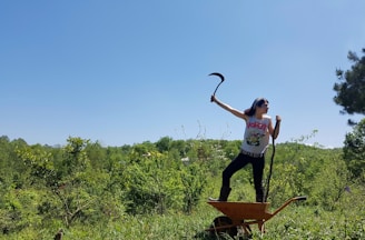 A person stands triumphantly on a wheelbarrow, holding a sickle in one hand and a stick in the other. The scene is set in a lush, green environment with dense foliage and a clear blue sky, suggesting a rural or forested area. The person is wearing a t-shirt and boots, indicating casual outdoor attire.