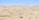 A vast desert landscape with rolling sand dunes and scattered sparse vegetation. Groups of people are walking and exploring the area, suggesting a tourist activity or hike. The clear blue sky enhances the arid beauty of the scenery.