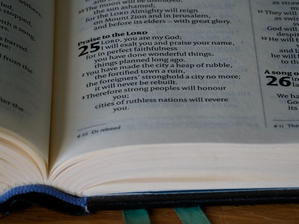 An open Bible is placed on a flat surface, displaying text from the Book of Isaiah. The visible verses include excerpts from chapter 25, which feature religious text written in both bold and regular fonts. A ribbon marker is slightly visible under the pages, and the lighting highlights the pages' texture.
