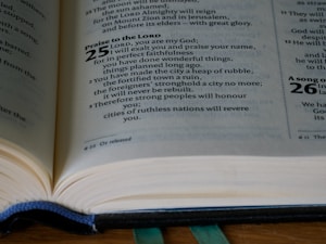 An open Bible is placed on a flat surface, displaying text from the Book of Isaiah. The visible verses include excerpts from chapter 25, which feature religious text written in both bold and regular fonts. A ribbon marker is slightly visible under the pages, and the lighting highlights the pages' texture.