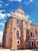 A well-preserved brick building featuring ornate architectural details. The structure has a rectangular shape with symmetrical windows and decorative elements on the facade, topped with an intricately designed, stepped gable. Adjacent to the building on the right is another structure with a pink facade, adding contrast to the scene.