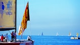 A sailboat with a red flag featuring intricate designs is in the foreground, occupied by several people engaging in various activities. The sailboat has a large yellow sail with text related to a nautical event in Venice. In the distance, several other sailboats are visible on the calm, blue sea under a clear sky. There is also a small dock structure with a yellow component to the right.