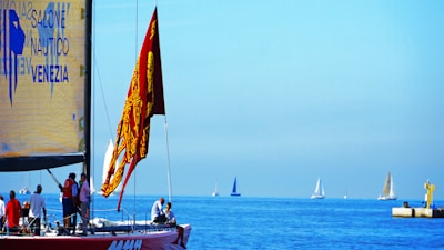 A sailboat with a red flag featuring intricate designs is in the foreground, occupied by several people engaging in various activities. The sailboat has a large yellow sail with text related to a nautical event in Venice. In the distance, several other sailboats are visible on the calm, blue sea under a clear sky. There is also a small dock structure with a yellow component to the right.