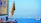 A sailboat with a red flag featuring intricate designs is in the foreground, occupied by several people engaging in various activities. The sailboat has a large yellow sail with text related to a nautical event in Venice. In the distance, several other sailboats are visible on the calm, blue sea under a clear sky. There is also a small dock structure with a yellow component to the right.