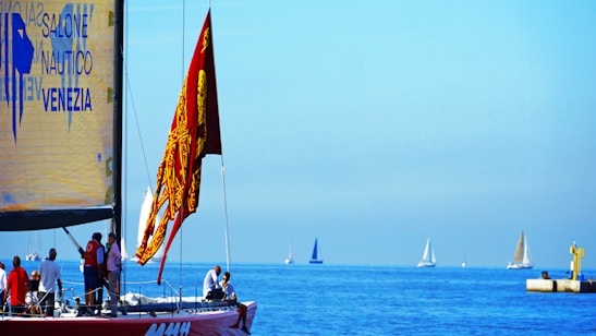 A sailboat with a red flag featuring intricate designs is in the foreground, occupied by several people engaging in various activities. The sailboat has a large yellow sail with text related to a nautical event in Venice. In the distance, several other sailboats are visible on the calm, blue sea under a clear sky. There is also a small dock structure with a yellow component to the right.