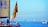 A sailboat with a red flag featuring intricate designs is in the foreground, occupied by several people engaging in various activities. The sailboat has a large yellow sail with text related to a nautical event in Venice. In the distance, several other sailboats are visible on the calm, blue sea under a clear sky. There is also a small dock structure with a yellow component to the right.