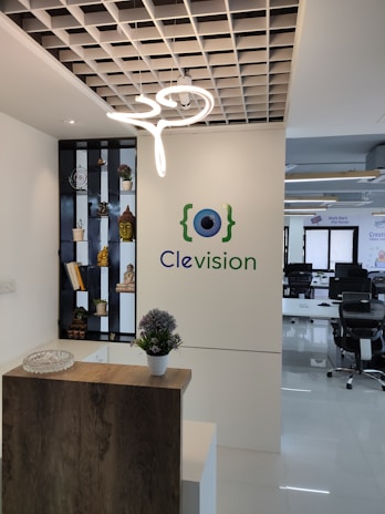 An office reception area with a decorative wooden counter and a potted plant on top. A modern, artistic light fixture hangs from a grid-like ceiling. A wall showcases the logo 'Clevision' with an eye symbol, and to the left, a shelf displays small plants and statues, including a golden Buddha head. In the background, several office chairs and desks can be seen.