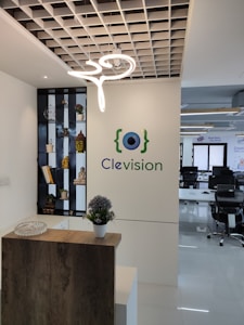 An office reception area with a decorative wooden counter and a potted plant on top. A modern, artistic light fixture hangs from a grid-like ceiling. A wall showcases the logo 'Clevision' with an eye symbol, and to the left, a shelf displays small plants and statues, including a golden Buddha head. In the background, several office chairs and desks can be seen.