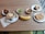 A wooden table is set with a variety of breakfast items. There's a cup of coffee with a heart-shaped design in the foam, placed on a saucer alongside a small packet and a napkin. Nearby, a small bowl contains a mix of pastries and dried fruits. Another bowl holds green grapes. A third bowl has pretzels, and there's a plate with a slice of bread topped with honey. A banana lies in the center, and a separate plate contains an assortment of cookies and a slice of cake.