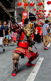 A person in a vibrant and elaborate costume strikes a dynamic pose on a street filled with people. Red lanterns are strung above the street, adding to the festive atmosphere. The costume features intricate designs with gold and red elements, including a striking mask and a pole or staff.