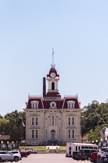 A historic courthouse with red roofs and a clock tower stands prominently in the center. The architecture is classic with multiple windows and decorative elements. American flags are seen on the building and there are several vehicles parked in the foreground. Trees are visible on both sides, providing a green backdrop against the clear blue sky.