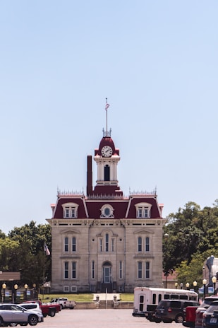 A historic courthouse with red roofs and a clock tower stands prominently in the center. The architecture is classic with multiple windows and decorative elements. American flags are seen on the building and there are several vehicles parked in the foreground. Trees are visible on both sides, providing a green backdrop against the clear blue sky.