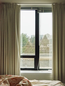 Curtains frame a window with a view of an overcast outdoor environment. The window overlooks rooftops, trees, and distant buildings under a cloudy sky. A bed with a floral-patterned blanket is visible in the foreground.