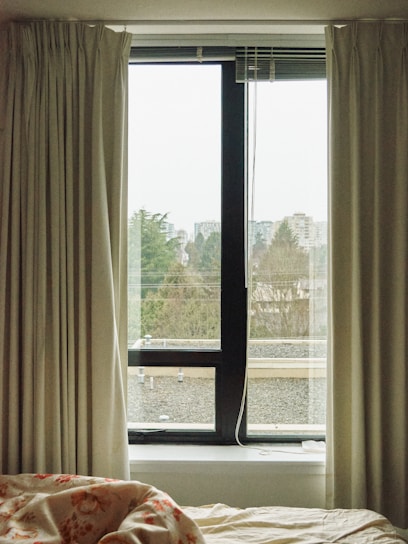 Curtains frame a window with a view of an overcast outdoor environment. The window overlooks rooftops, trees, and distant buildings under a cloudy sky. A bed with a floral-patterned blanket is visible in the foreground.
