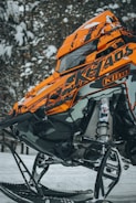 A close-up of a brightly colored snowmobile with an orange and black design. The vehicle is parked on snowy terrain surrounded by snow-covered trees. The detailed view highlights the mechanical components and sleek design features.