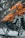 A close-up of a brightly colored snowmobile with an orange and black design. The vehicle is parked on snowy terrain surrounded by snow-covered trees. The detailed view highlights the mechanical components and sleek design features.