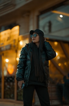 A person wearing a black puffer jacket, sunglasses, and a cap stands confidently in an urban environment with warm lighting and blurred background.