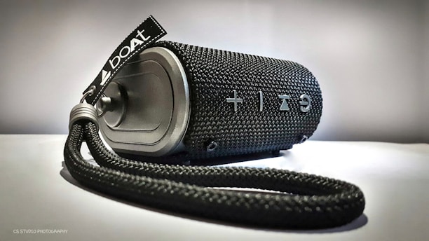 A cylindrical portable speaker with a black woven texture and a carrying strap attached. It features prominent white control symbols, including a plus and minus sign for volume adjustment. The brand name 'boat' is displayed on a tag attached to the strap.