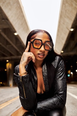 A person with large, geometric sunglasses poses confidently under an overpass. The individual is wearing a textured black outfit and has tattoos on their hand. The setting is urban, with concrete structures and dim lighting in the background.