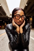 A woman wearing oversized geometric sunglasses and a textured black jacket is posing with her hands framing her face. The background is a dimly lit urban setting, possibly under a bridge or overpass.