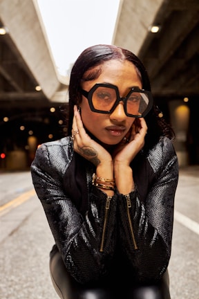 A woman wearing oversized geometric sunglasses and a textured black jacket is posing with her hands framing her face. The background is a dimly lit urban setting, possibly under a bridge or overpass.