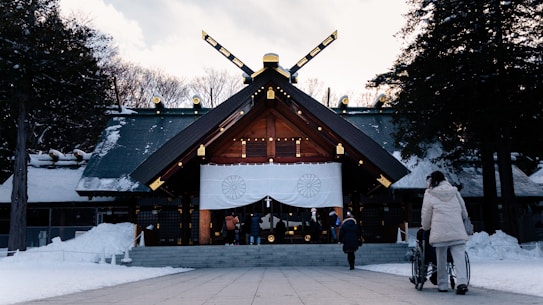 A traditional Japanese shrine with a large entrance draped in white banners featuring symbolic emblems. The shrine is set in a snowy landscape surrounded by trees. Visitors are seen approaching the entrance, some dressed warmly for the cold weather. The roof is adorned with gold accents and unique architectural features.
