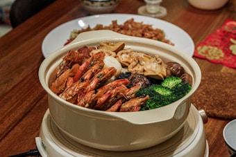 A pot filled with various ingredients including prawns, broccoli, mushrooms, and seafood is placed on a wooden table. The dish is served in a white ceramic pot, and there are additional plates and festive decorations in the background.