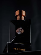 A luxurious black bottle of Versace perfume with a metallic bronze cap, featuring a Medusa emblem in the center. The brand name is prominently displayed in gold lettering on the base of the bottle.