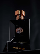 A luxurious black bottle of Versace perfume with a metallic bronze cap, featuring a Medusa emblem in the center. The brand name is prominently displayed in gold lettering on the base of the bottle.