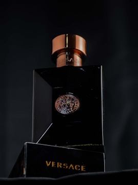 A luxurious black bottle of Versace perfume with a metallic bronze cap, featuring a Medusa emblem in the center. The brand name is prominently displayed in gold lettering on the base of the bottle.