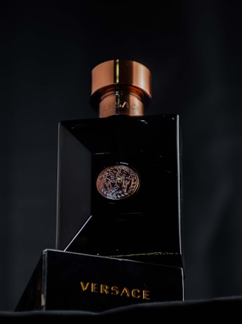 A luxurious black bottle of Versace perfume with a metallic bronze cap, featuring a Medusa emblem in the center. The brand name is prominently displayed in gold lettering on the base of the bottle.