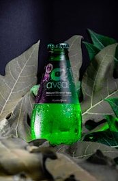 A green glass bottle of Avşar natural mineral water is positioned upright among large, deep green leaves. Water droplets cover the surface of the bottle, giving it a fresh, chilled appearance. The label is predominantly dark with white text.