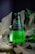 A green glass bottle of Avşar natural mineral water is positioned upright among large, deep green leaves. Water droplets cover the surface of the bottle, giving it a fresh, chilled appearance. The label is predominantly dark with white text.