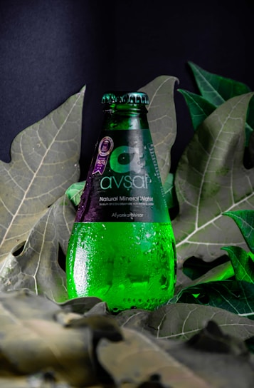 A green glass bottle of Avşar natural mineral water is positioned upright among large, deep green leaves. Water droplets cover the surface of the bottle, giving it a fresh, chilled appearance. The label is predominantly dark with white text.