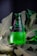 A green glass bottle of Avşar natural mineral water is positioned upright among large, deep green leaves. Water droplets cover the surface of the bottle, giving it a fresh, chilled appearance. The label is predominantly dark with white text.