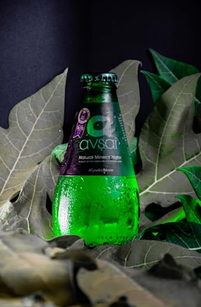A green glass bottle of Avşar natural mineral water is positioned upright among large, deep green leaves. Water droplets cover the surface of the bottle, giving it a fresh, chilled appearance. The label is predominantly dark with white text.