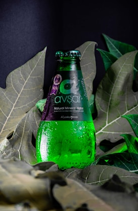 A green glass bottle of Avşar natural mineral water is positioned upright among large, deep green leaves. Water droplets cover the surface of the bottle, giving it a fresh, chilled appearance. The label is predominantly dark with white text.