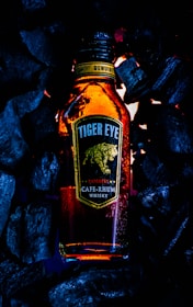 A bottle labeled 'Tiger Eye CAFÉ-RHUM Whisky' surrounded by dark charcoal pieces emits a warm, amber glow. The label features a stylized image of a tiger in motion, and there are water droplets on the bottle suggesting condensation.