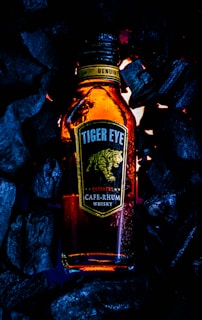 A bottle labeled 'Tiger Eye CAFÉ-RHUM Whisky' surrounded by dark charcoal pieces emits a warm, amber glow. The label features a stylized image of a tiger in motion, and there are water droplets on the bottle suggesting condensation.