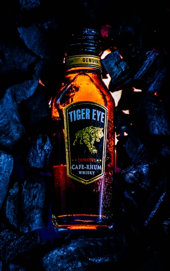 A bottle labeled 'Tiger Eye CAF&Eacute;-RHUM Whisky' surrounded by dark charcoal pieces emits a warm, amber glow. The label features a stylized image of a tiger in motion, and there are water droplets on the bottle suggesting condensation.