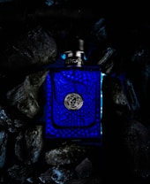 A dark, moody image displaying a blue bottle with a silver cap, featuring intricate textured patterns on its surface. The bottle is situated among dark, irregularly shaped rocks or charcoal, creating a striking contrast with the vibrant blue color of the bottle.