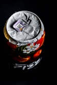 A close-up view of the top of a can with a crumpled silver foil lid, featuring a colorful design of white flowers and oranges on a red background. A small QR code sticker with a flag detail is placed on the lid.