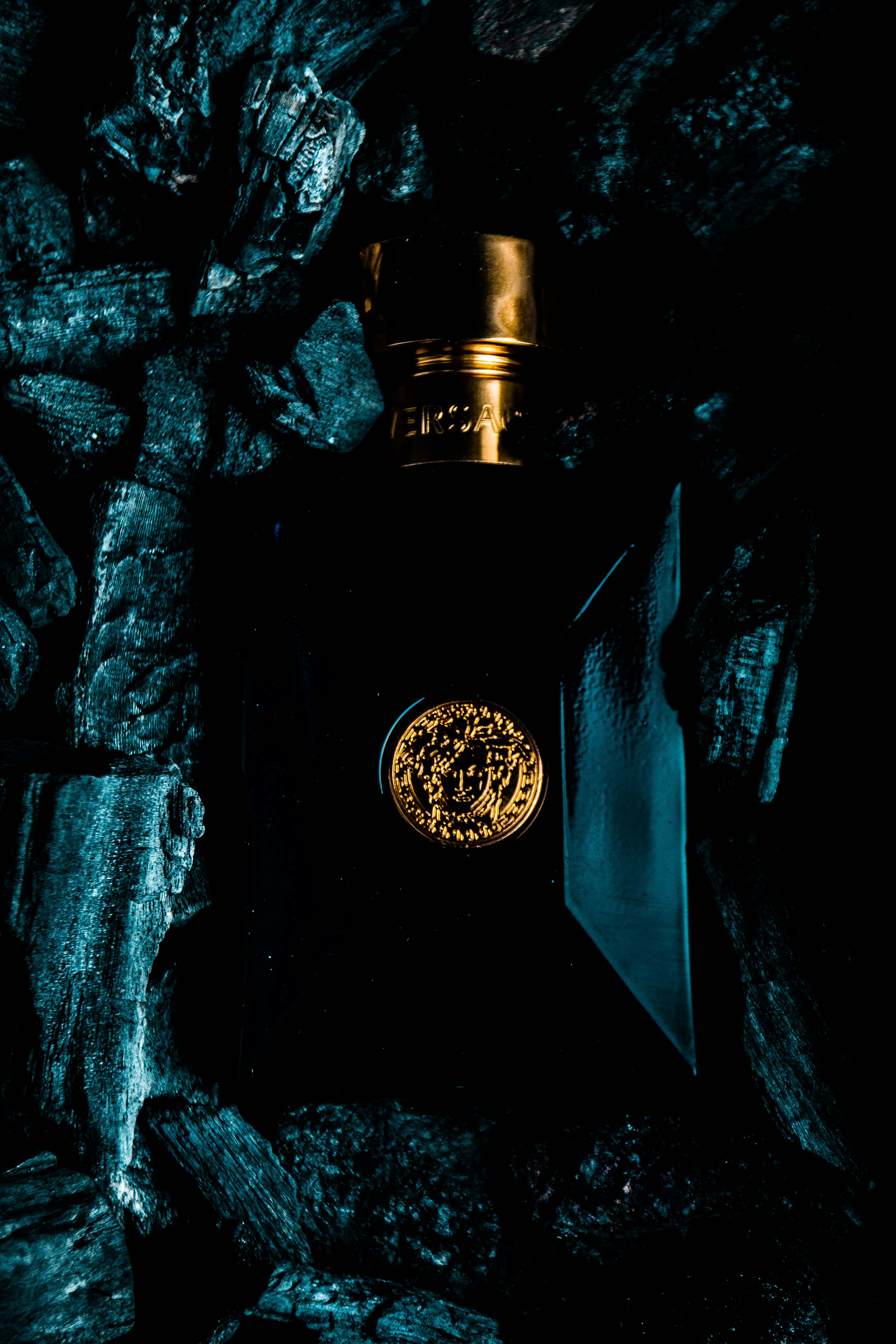 A luxurious perfume bottle with a gold cap and emblem is surrounded by dark, textured charcoal pieces. The bottle's design features elegant lines and a glossy finish, reflecting ambient light.