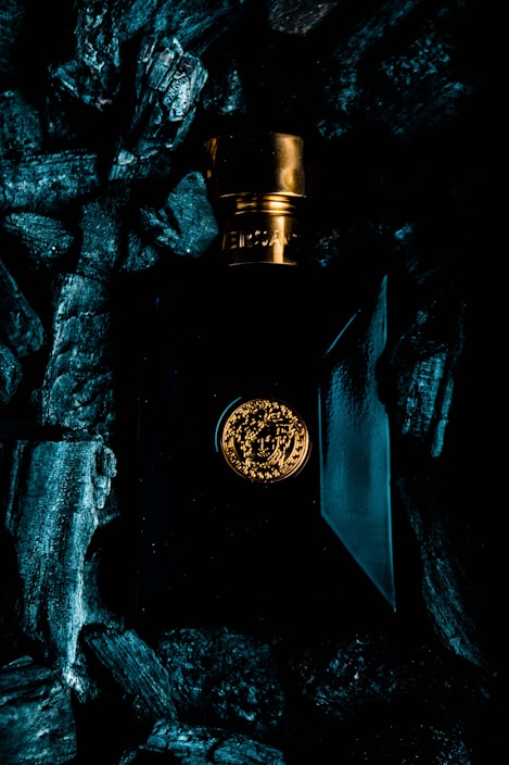 A luxurious perfume bottle with a gold cap and emblem is surrounded by dark, textured charcoal pieces. The bottle's design features elegant lines and a glossy finish, reflecting ambient light.