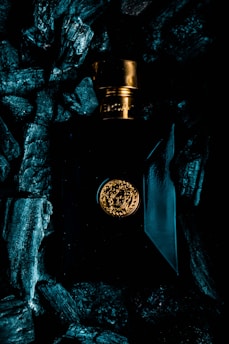 A luxurious perfume bottle with a gold cap and emblem is surrounded by dark, textured charcoal pieces. The bottle's design features elegant lines and a glossy finish, reflecting ambient light.