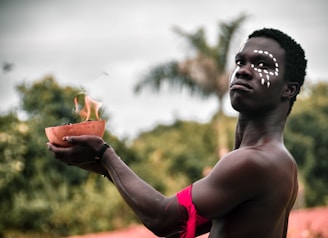 A person with white facial paint holds a burning bowl, appearing contemplative against a backdrop of blurred greenery. The red paint or cloth on the arm adds contrast, and palm trees are faintly visible in the background.