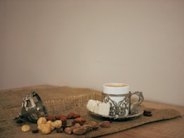 A decorative coffee cup with an ornate metal holder is placed on a rustic burlap fabric. Next to it are pieces of white Turkish delight and an array of mixed nuts, including hazelnuts, almonds, and pistachios. An overturned metal cup lies beside them, contributing to the vintage and cozy atmosphere.