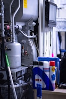 A cluttered industrial or mechanical workspace with various machines and equipment. There is a complex apparatus with pipes and metallic components, alongside a desk holding cleaning supplies, sprays, and folders. A digital screen is mounted on the wall, possibly for monitoring or control purposes.
